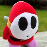Close-up of red Shy Guy plush showing white mask and black embroidered facial details
