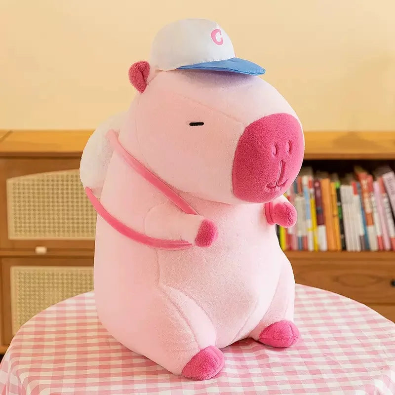 Pink capybara plush wearing a blue-and-white cap and small backpack with pink straps